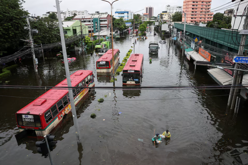 Illustrative image of severe flooding in Bangkok, Thailand, showing submerged vehicles, flooded streets and residents wading through water under dark storm clouds.
